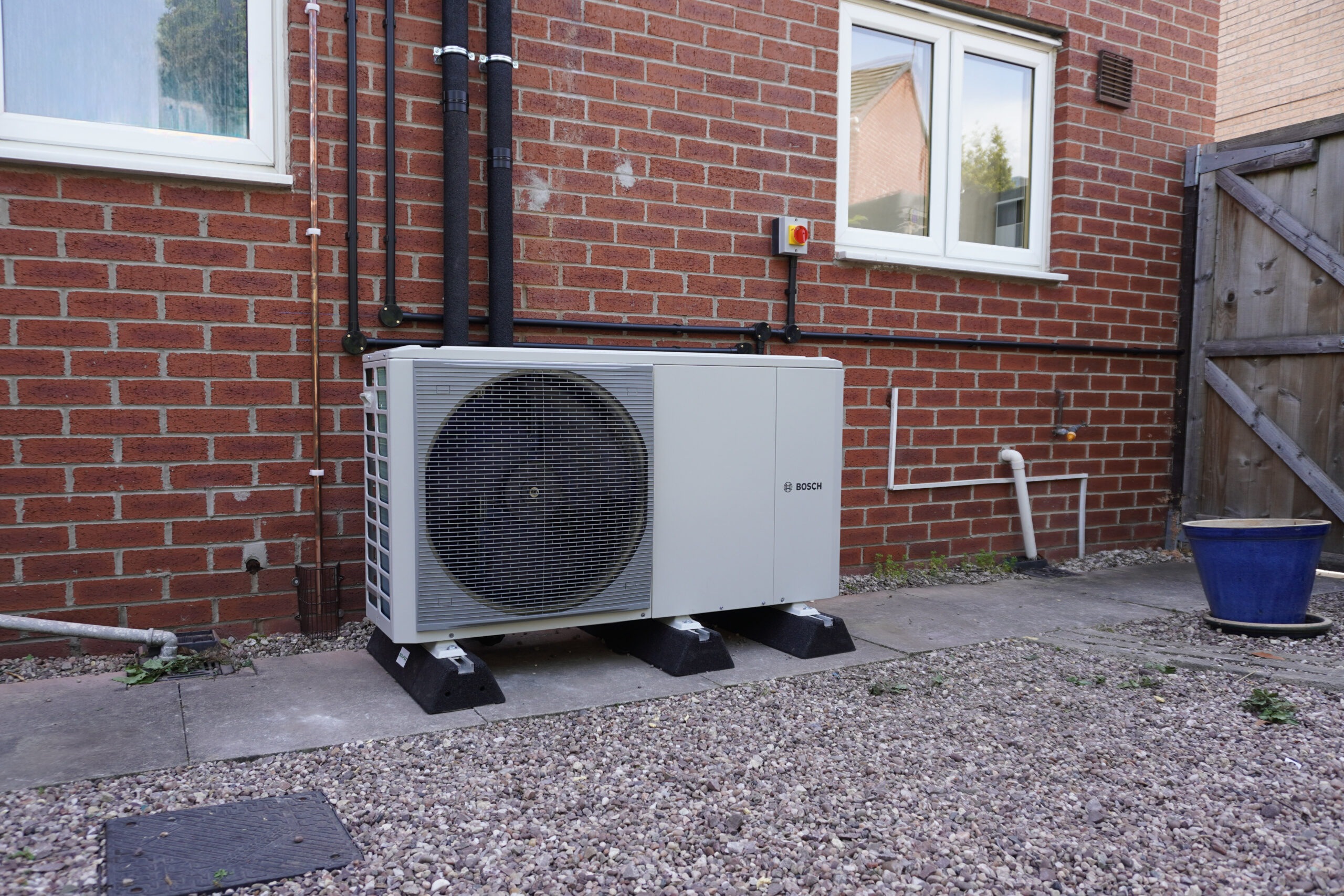 A Bosch air source heat pump unit installed on a concrete patio against a red brick exterior wall of a house, featuring insulated black pipework running up the wall and a blue plant pot nearby.